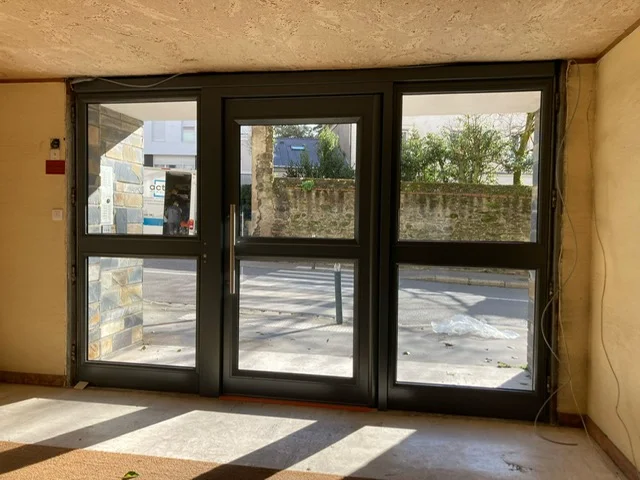 Porte d'entrée d'une résidence située en ville, entourée de deux fenêtre toute hauteur favorisant l'entrée de lumière dans la résidence.