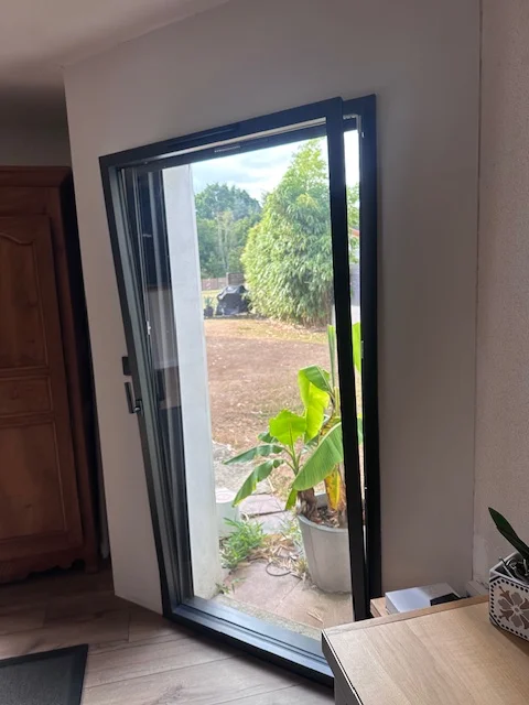 Porte-fenêtre aluminium moderne noire entrouverte sur un jardin. Un chantier réalisé par Actiba Menuiseries.